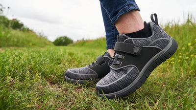 Dark grey walking shoes with wide hook-and-loop strap closure and textured knit upper, on-model outdoor shot on grass showing cushioned collar padding and black rubber outsole