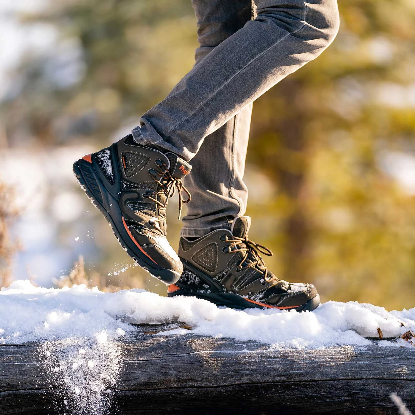 Black and tan hiking boots with orange accent trim and lace hooks, on-model action shot walking on snowy log displaying high-top ankle support and lugged rubber outsole