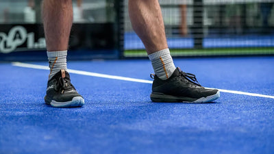Black tennis shoes with white midsole and lace-up closure, on-model action shot on blue court surface showing low-top silhouette and herringbone outsole pattern