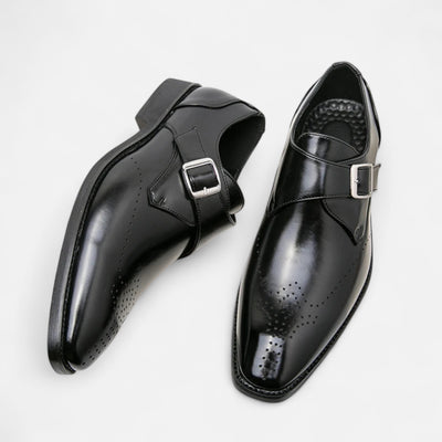 Black leather single monk strap dress shoes with brogue perforation detail and silver buckle closure, overhead paired view showing medallion toe cap and leather sole construction