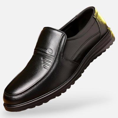 Black grain leather slip-on shoe with decorative stitching detail and metal badge embellishment, angled view showing thick ridged rubber comfort sole and yellow heel tab
