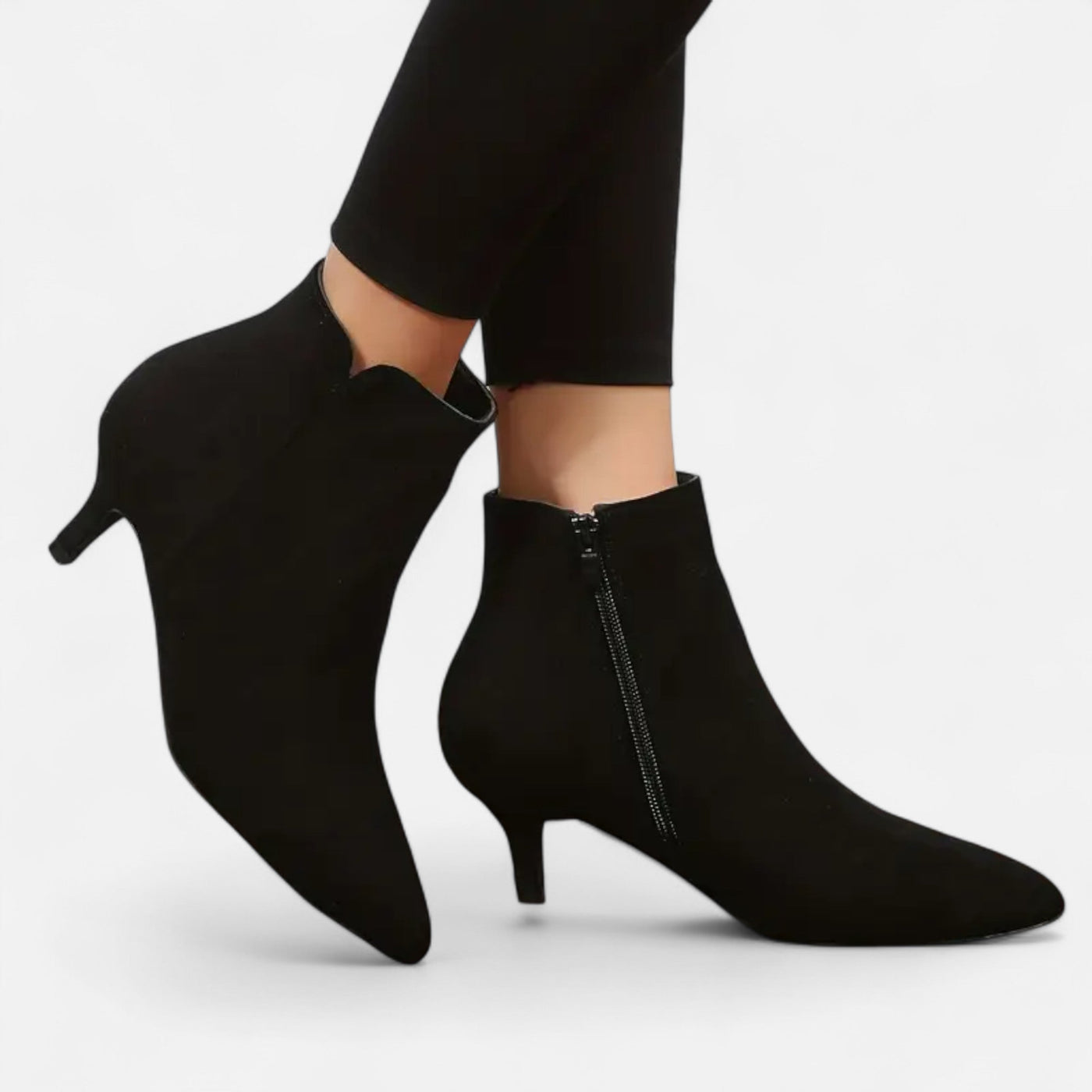 Black suede ankle booties with exposed side zipper fastening and kitten heel, on-model worn view displaying pointed toe shape and slim heel construction