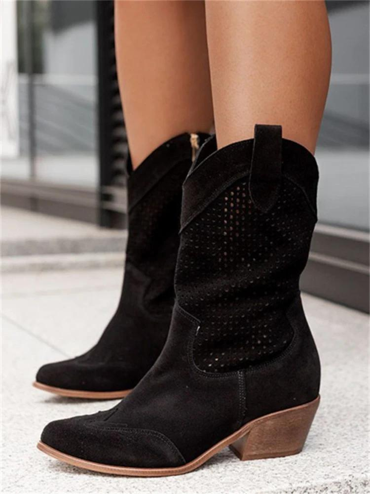 Black suede cowboy boots with decorative cutout perforation pattern and contrast wood heel, on-model front angle displaying pointed toe and mid-calf shaft with pull tabs