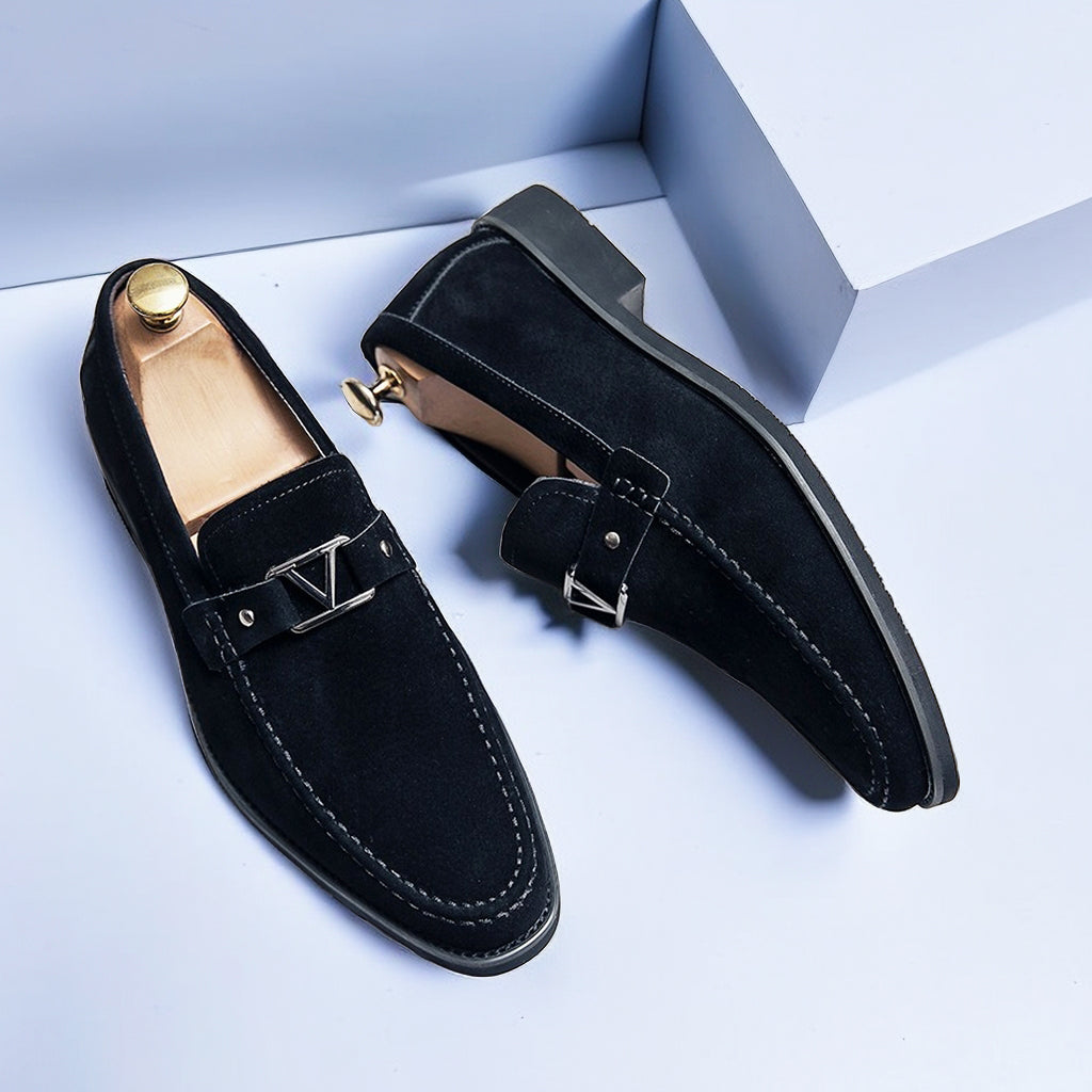Black suede loafers with decorative metal buckle hardware and contrast white topstitching, paired flat lay view with wooden shoe trees showing moc-toe construction and rubber sole
