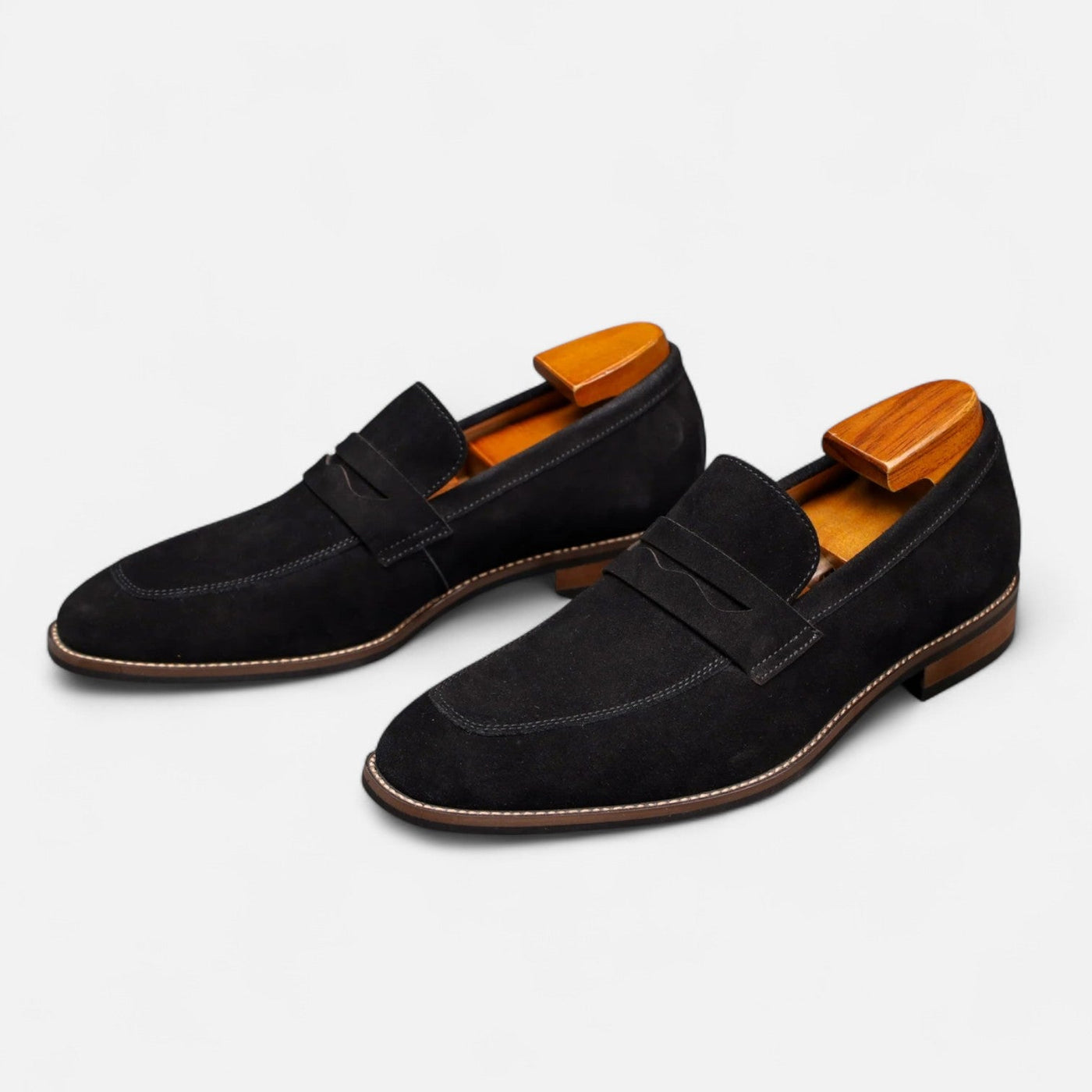 Black suede slip-on loafers with tonal penny keeper and grey contrast topstitching, paired perspective showing brown leather sole construction with wooden shoe forms