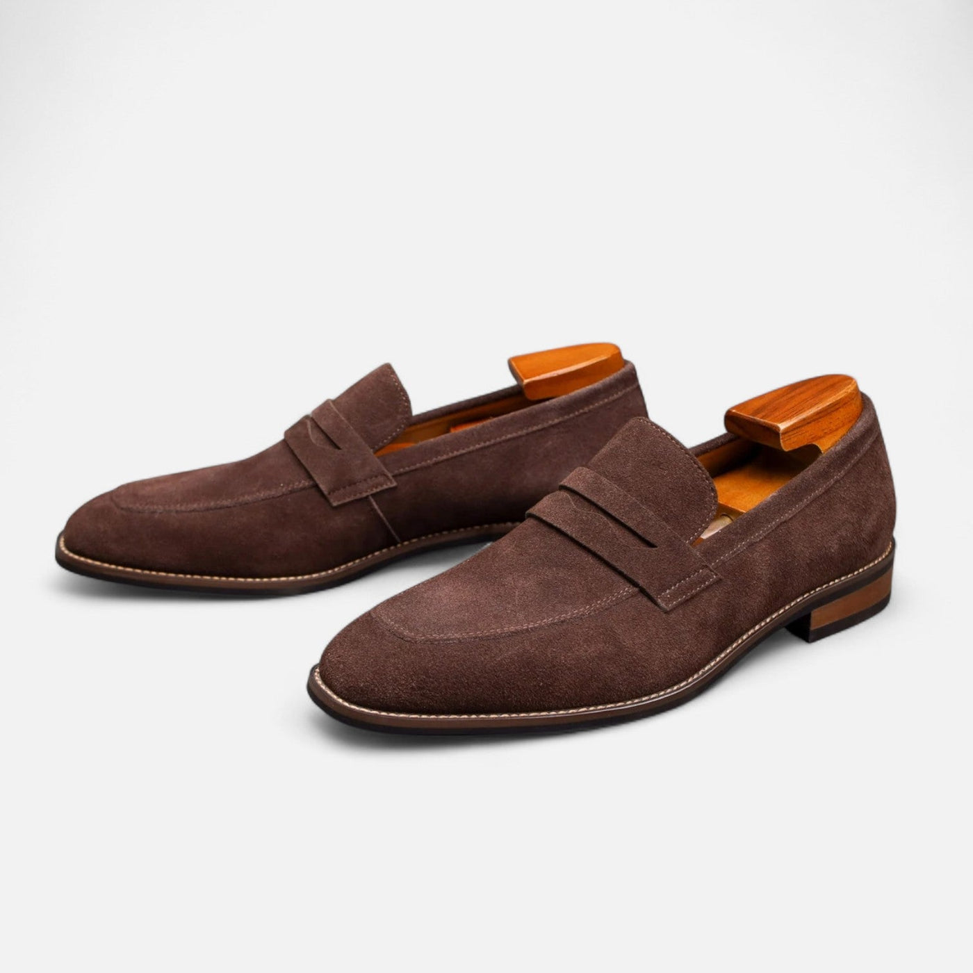 Brown suede penny loafer slip-on shoes with contrast white topstitching and leather sole, paired three-quarter view with cedar shoe trees inserted